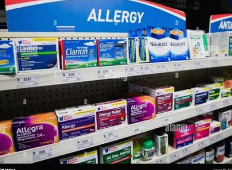 Pharmacist retrieving a box of allergy medication from a shelf behind the pharmacy counter while a customer waits with identification in hand