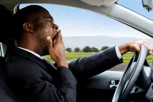 A driver in a car interior with a sleepy expression, hands on the steering wheel