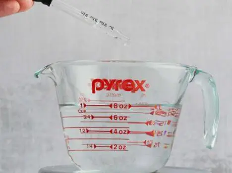 A person measuring a dose of oral liquid laxative into a small cup next to a full glass of water.