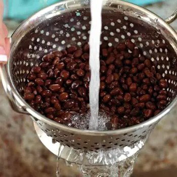 Canned beans being rinsed under running water in a metal colander in a kitchen sink