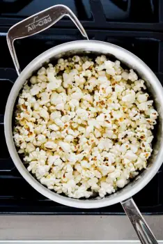 A metal pot on a stovetop with the lid slightly open, revealing freshly popped white popcorn inside.